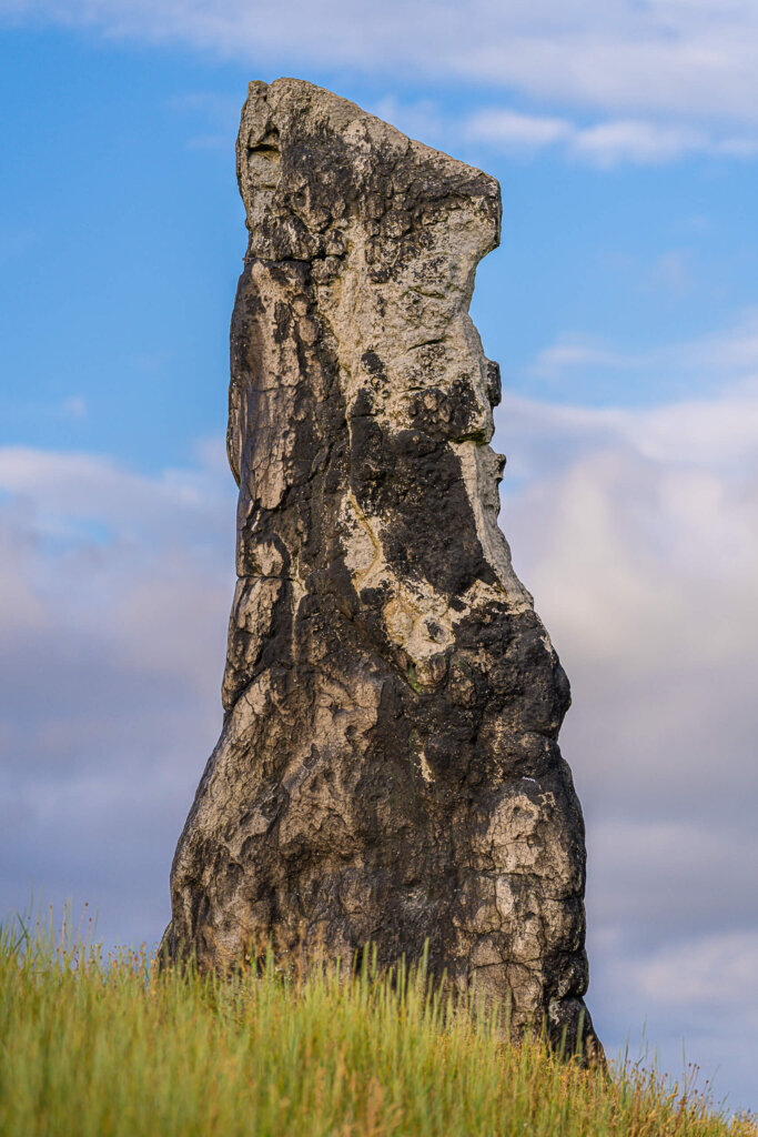 Menhir Steinerne Jungfrau in Dölau: Ein hoch aufragender, verwitterter Steinmonolith auf einem Grashügel vor blauem Wolkenhimmel.