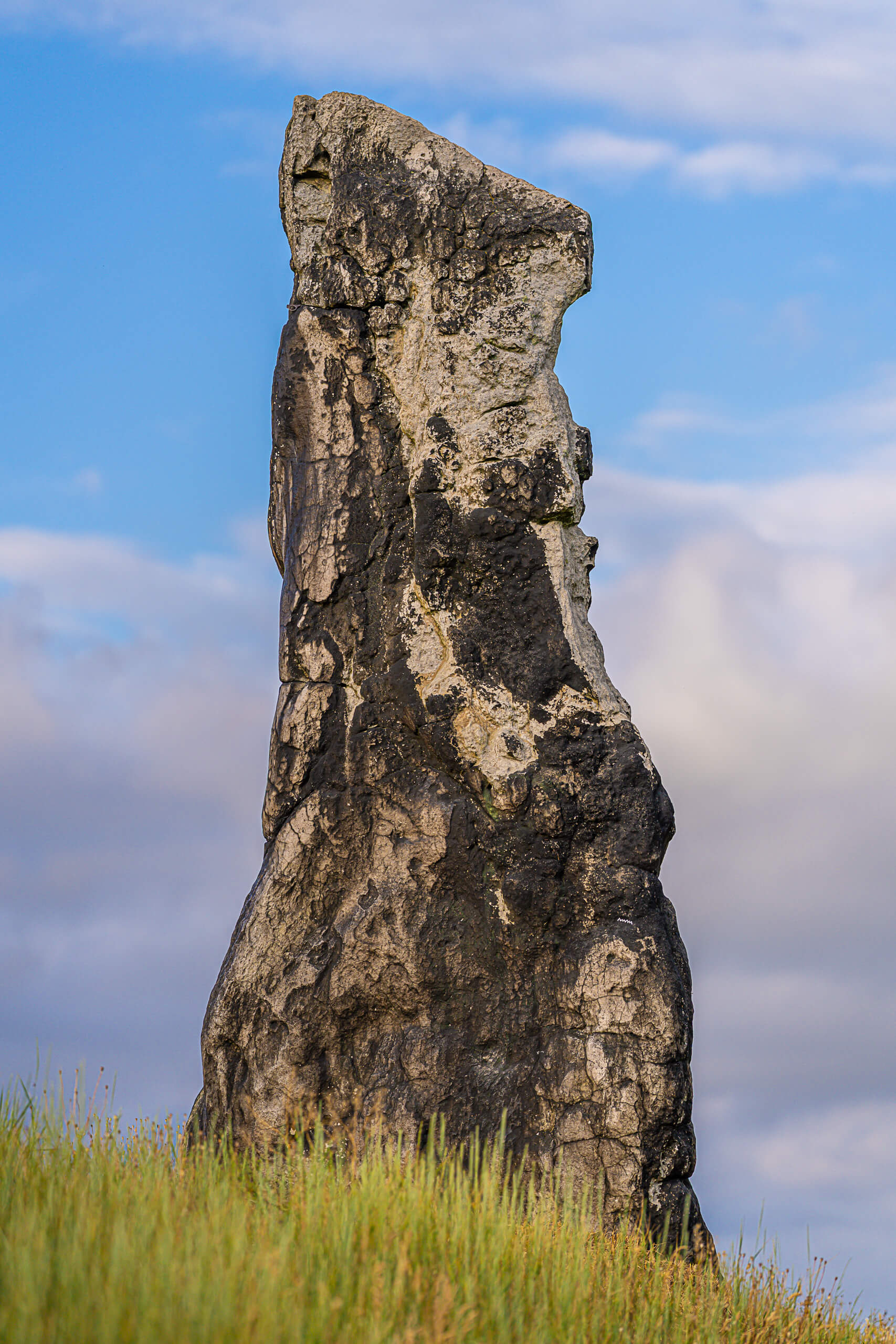 Menhir Steinerne Jungfrau in Dölau: Ein hoch aufragender, verwitterter Steinmonolith auf einem Grashügel vor blauem Wolkenhimmel.